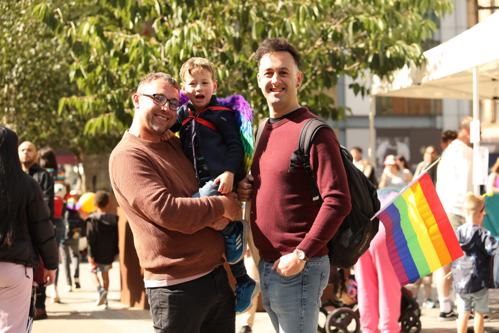 Two LGBT+ parents smile at the camera and embrace as they hold their child in their arms at Family Pride MCR.