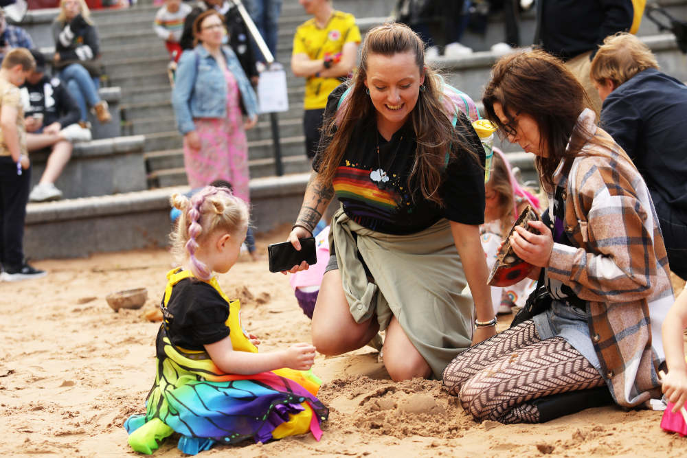 Two LGBT+ parents play with their child, who is dressed in a rainbow butterfly costume, in a sand pit at Family Pride MCR.